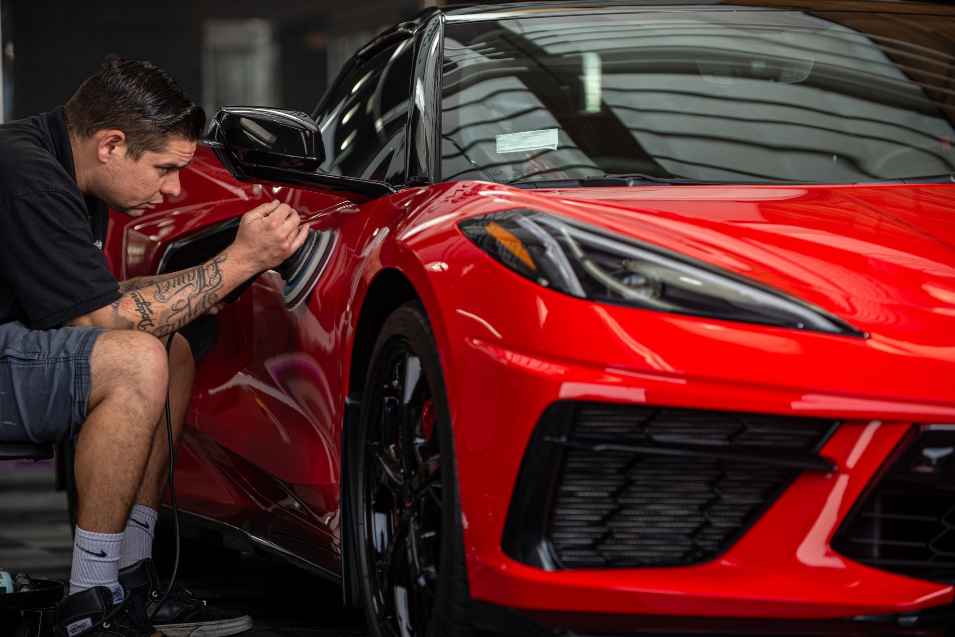 Man polishing a red sports car. He is crouching with a polisher in hand, working on the car's side.