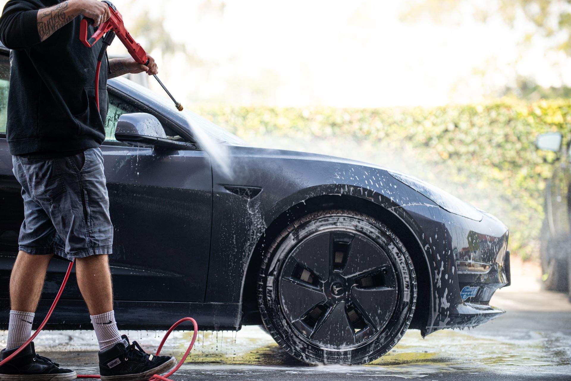Person washing a dark gray Tesla with a pressure washer, soap suds visible.