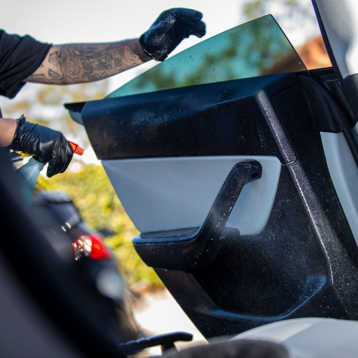 Person with tattooed arm applying tint film to a car window. Spray bottle in hand, black gloves.
