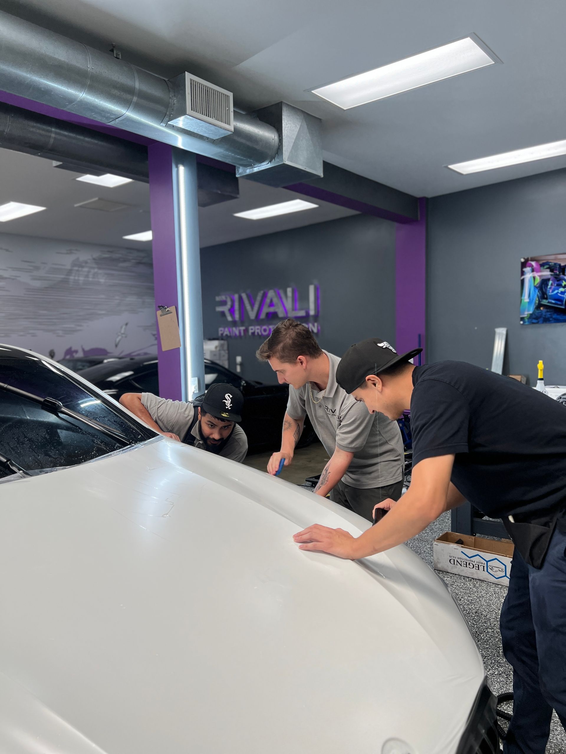 Three people working on the hood of a white car inside a shop. The shop has a gray and purple color scheme.