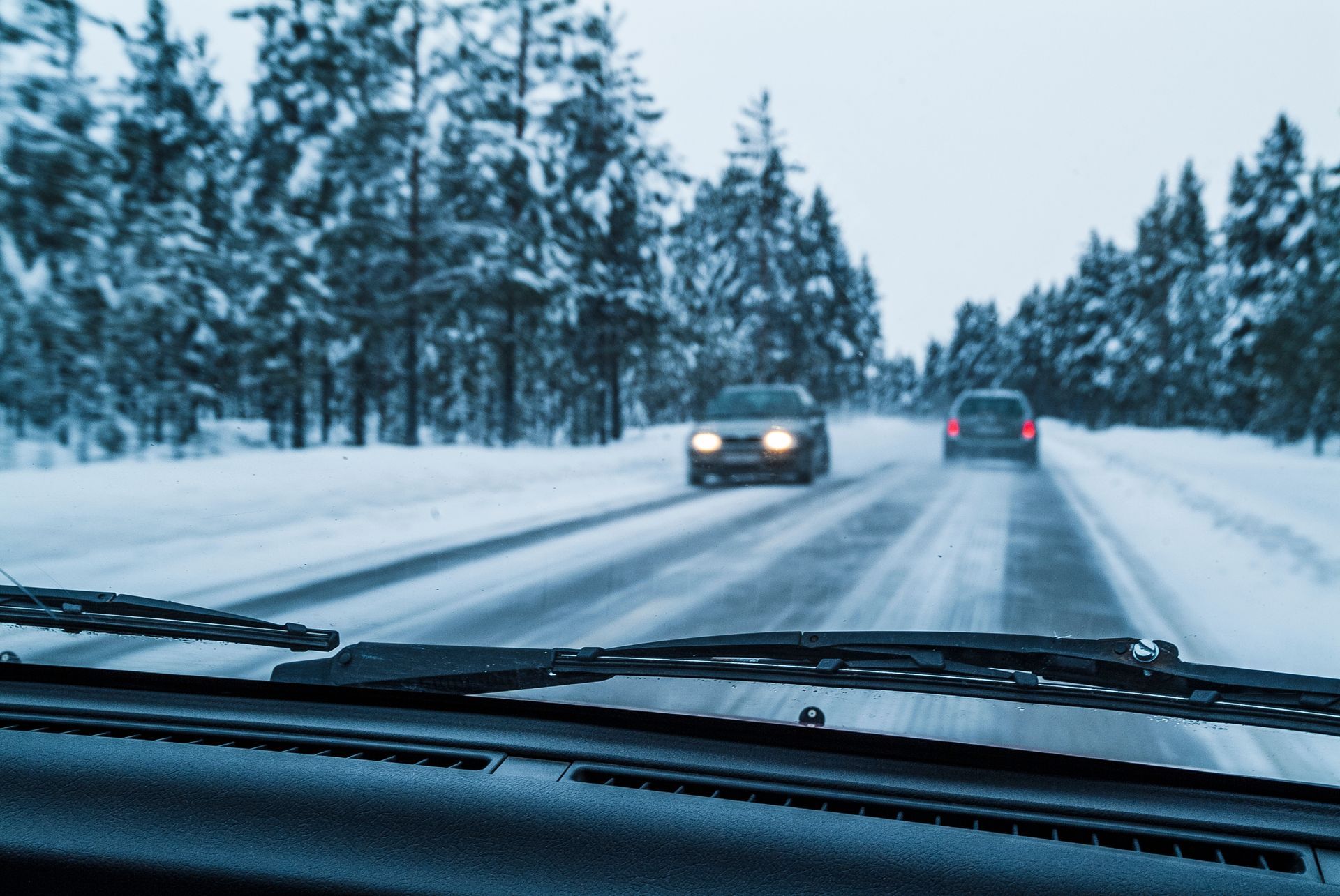 Snowy road seen from car windshield, with two vehicles ahead in wintry forest landscape.