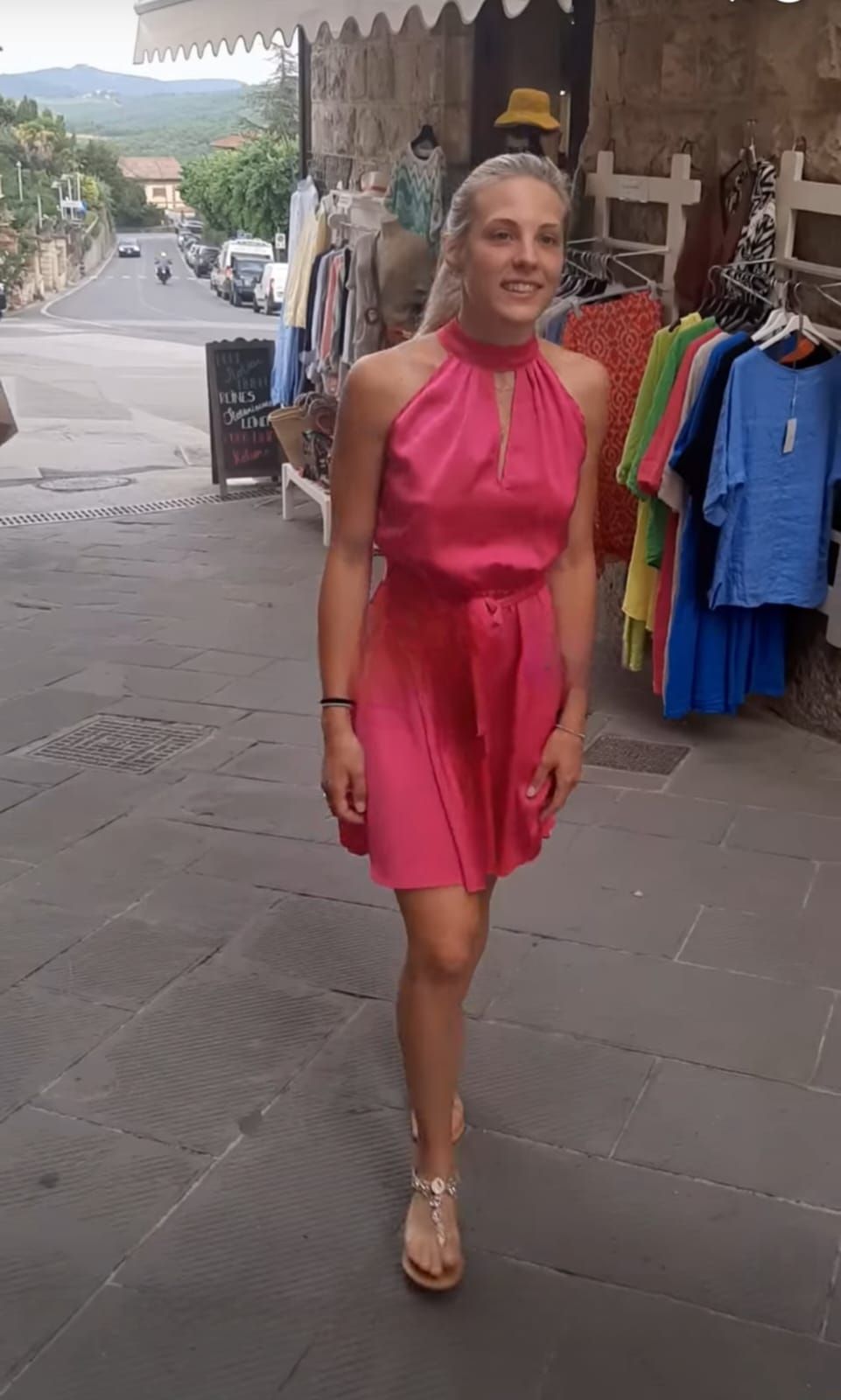 Donna con un vestito rosa acceso cammina su un marciapiede davanti alle vetrine dei negozi all'aperto