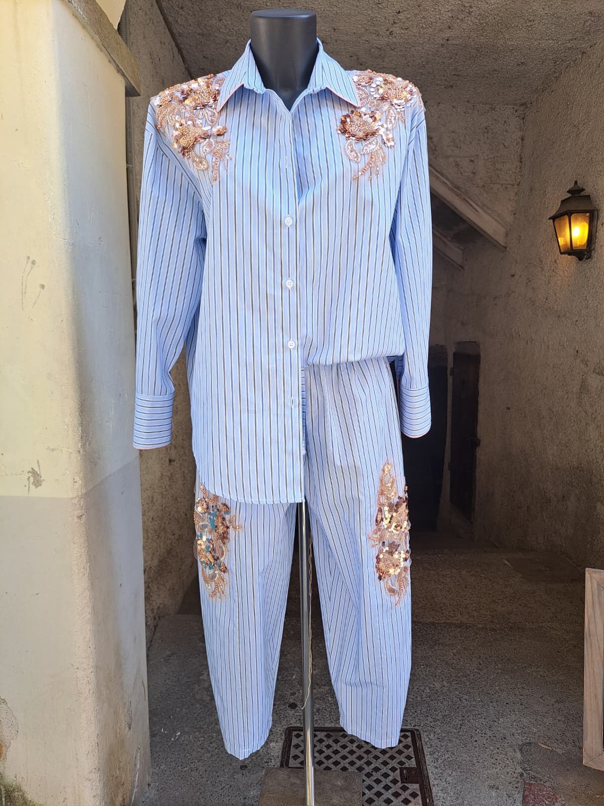 Completo composto da camicia e pantaloni a righe bianche con applicazioni floreali color pesca, esposto su un manichino in un corridoio.