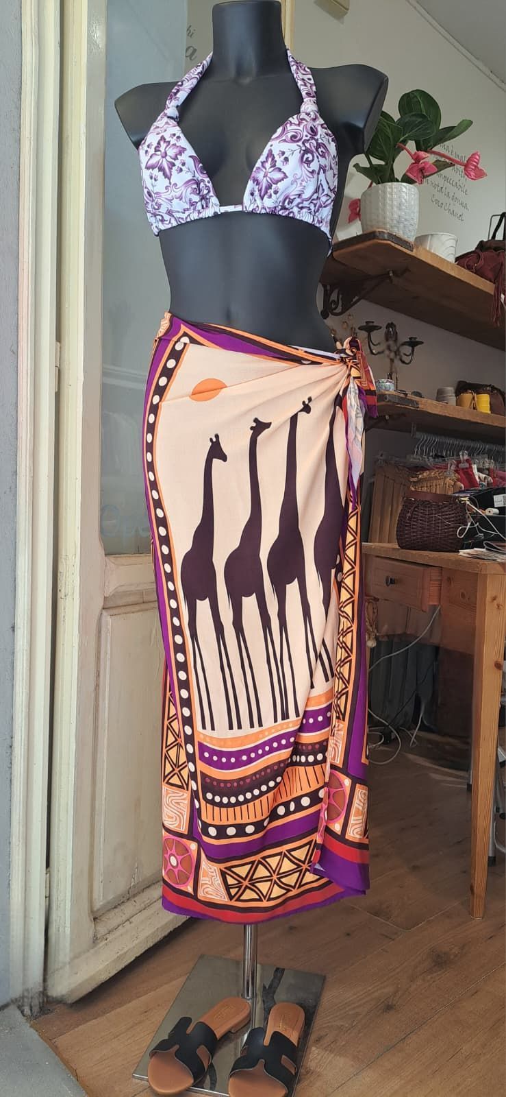 Manichino che indossa un top bikini a fiori e una gonna a portafoglio fantasia, in piedi in un ambiente interno con le scarpe nelle vicinanze.