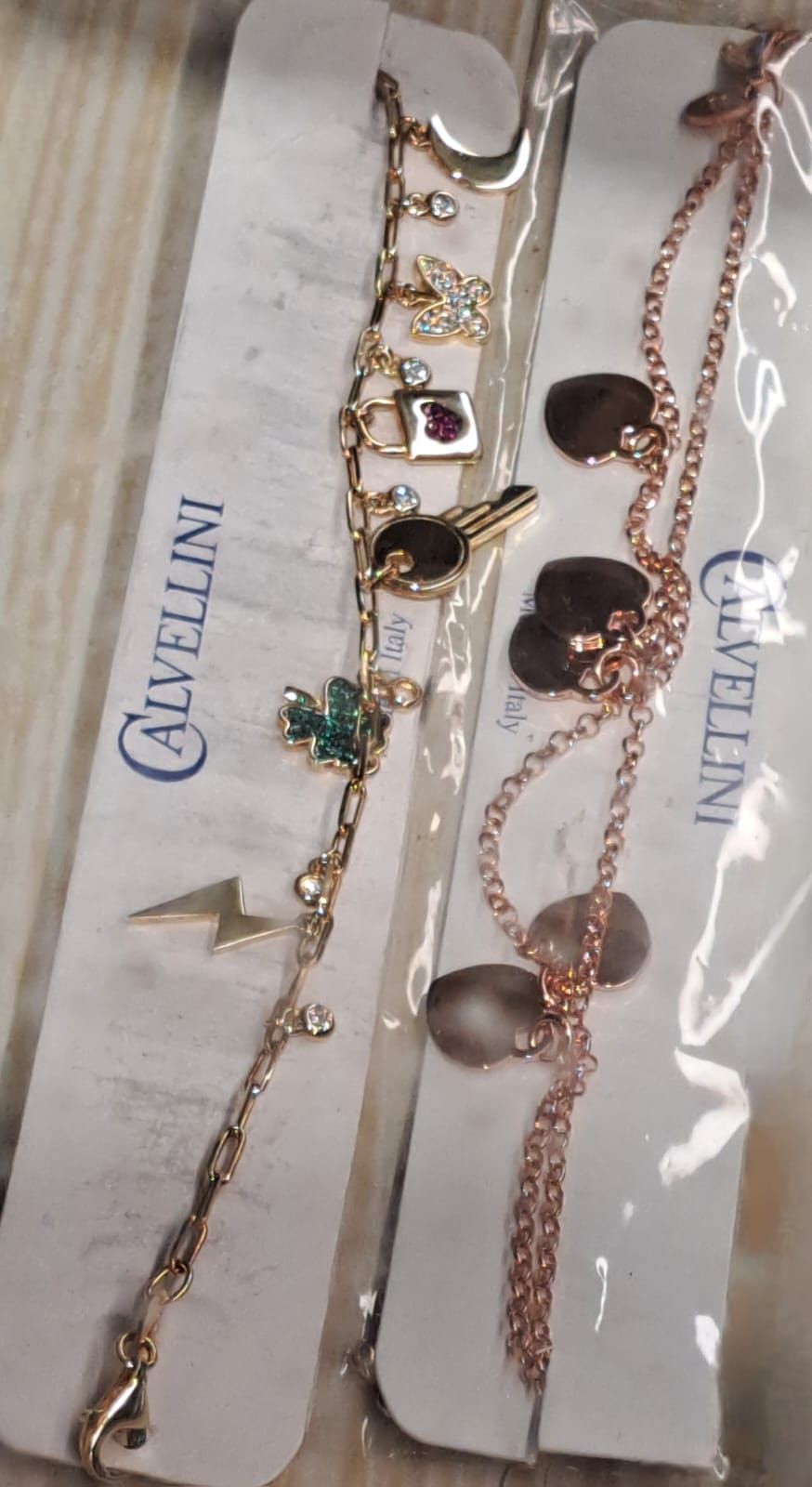 Due braccialetti con ciondoli color oro su cartoncini bianchi, uno con ciondoli a forma di cuore e uno con gemme colorate.