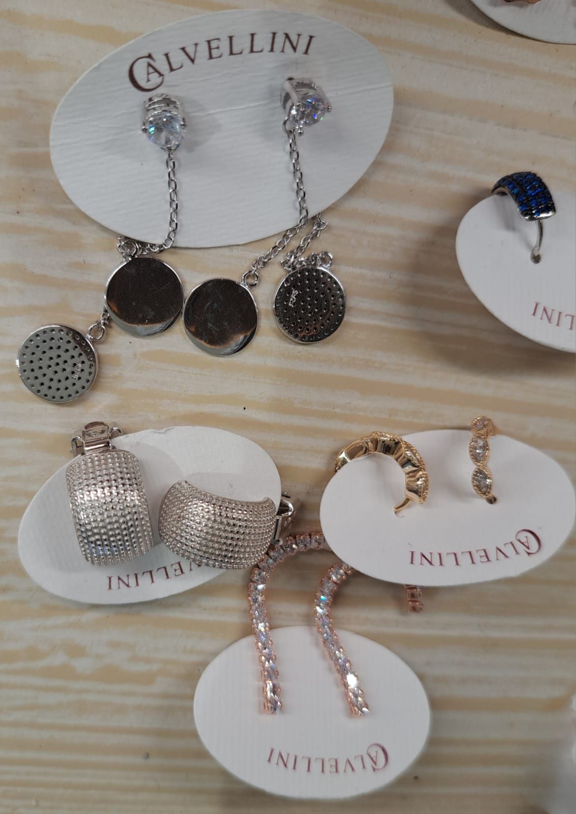 Assortimento di orecchini pendenti in argento e oro esposti su cartoncini bianchi con logo su una superficie di legno.