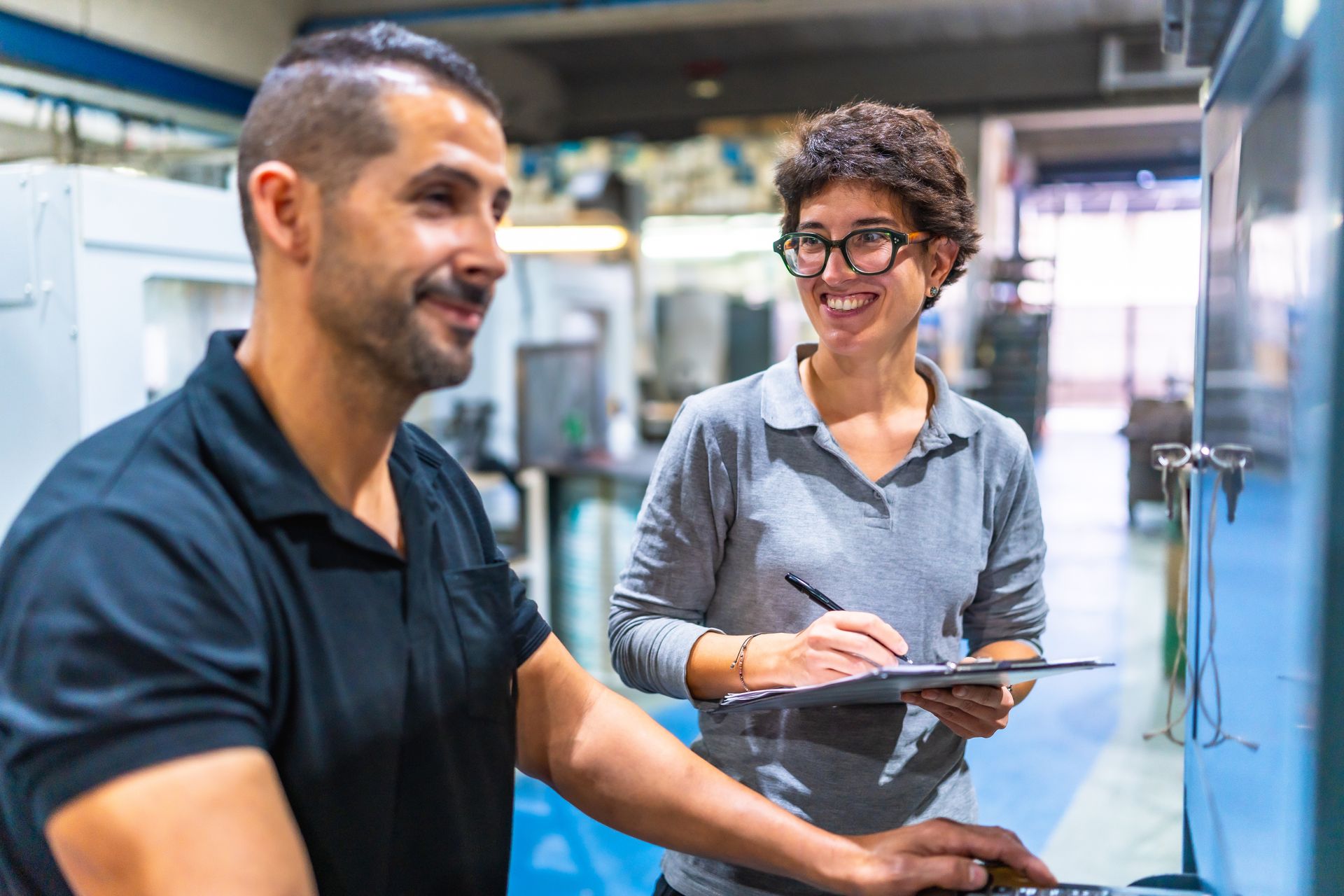 A smiling person in a gray polo writes on a clipboard while standing next to a colleague operating machinery in a factory.
