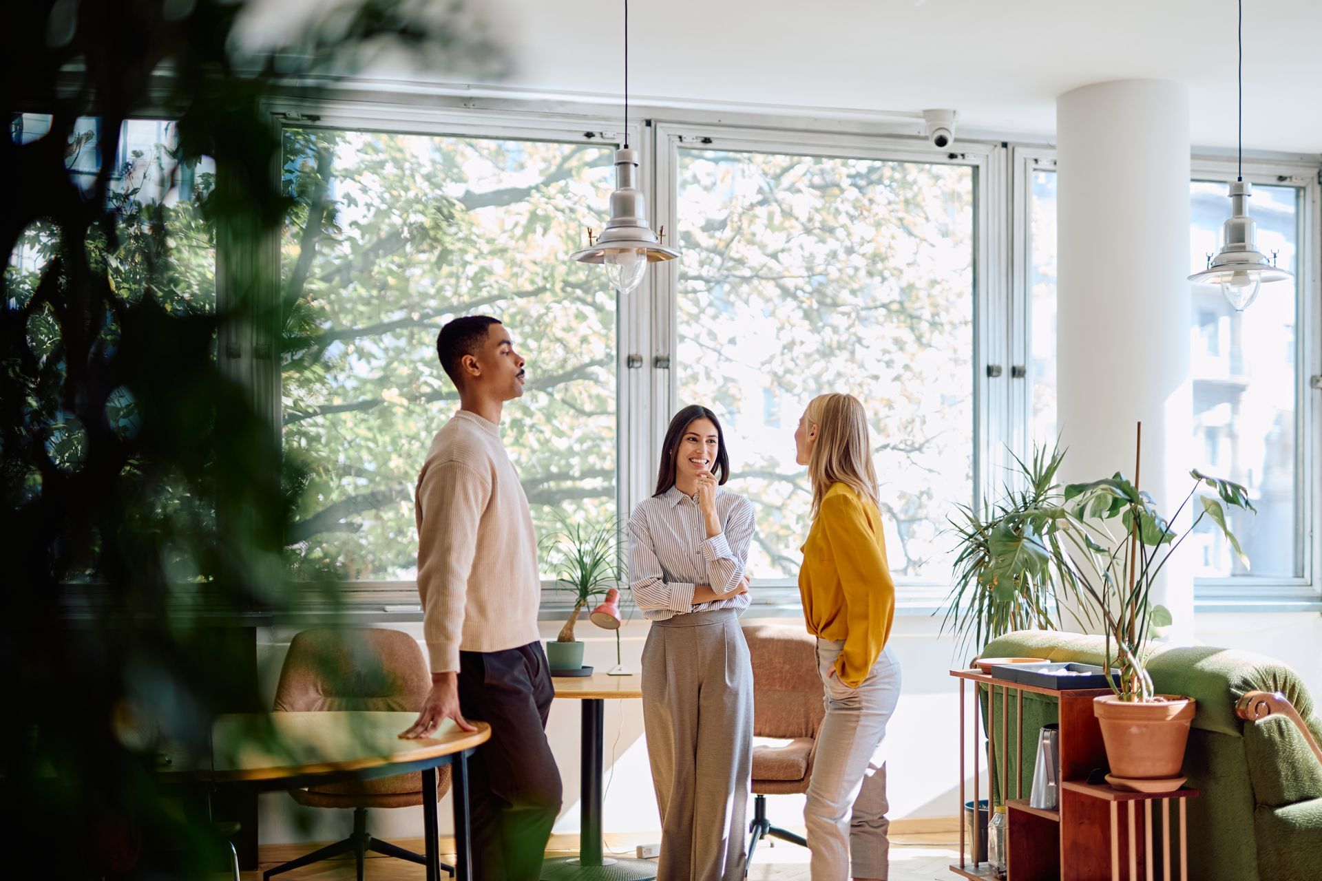 Three people talk together in a bright, modern office space with large windows, tables, chairs, and potted plants.