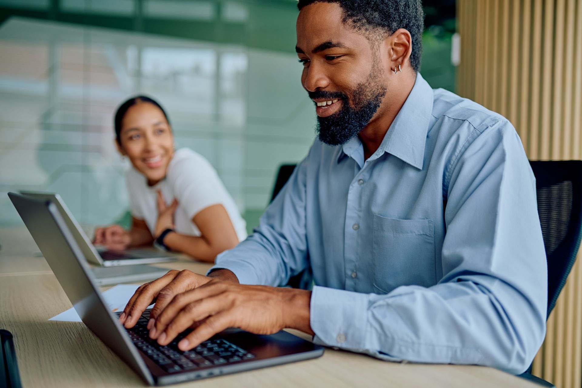 A person types on a laptop at a desk in an office, while another person sits in the background, smiling toward them.