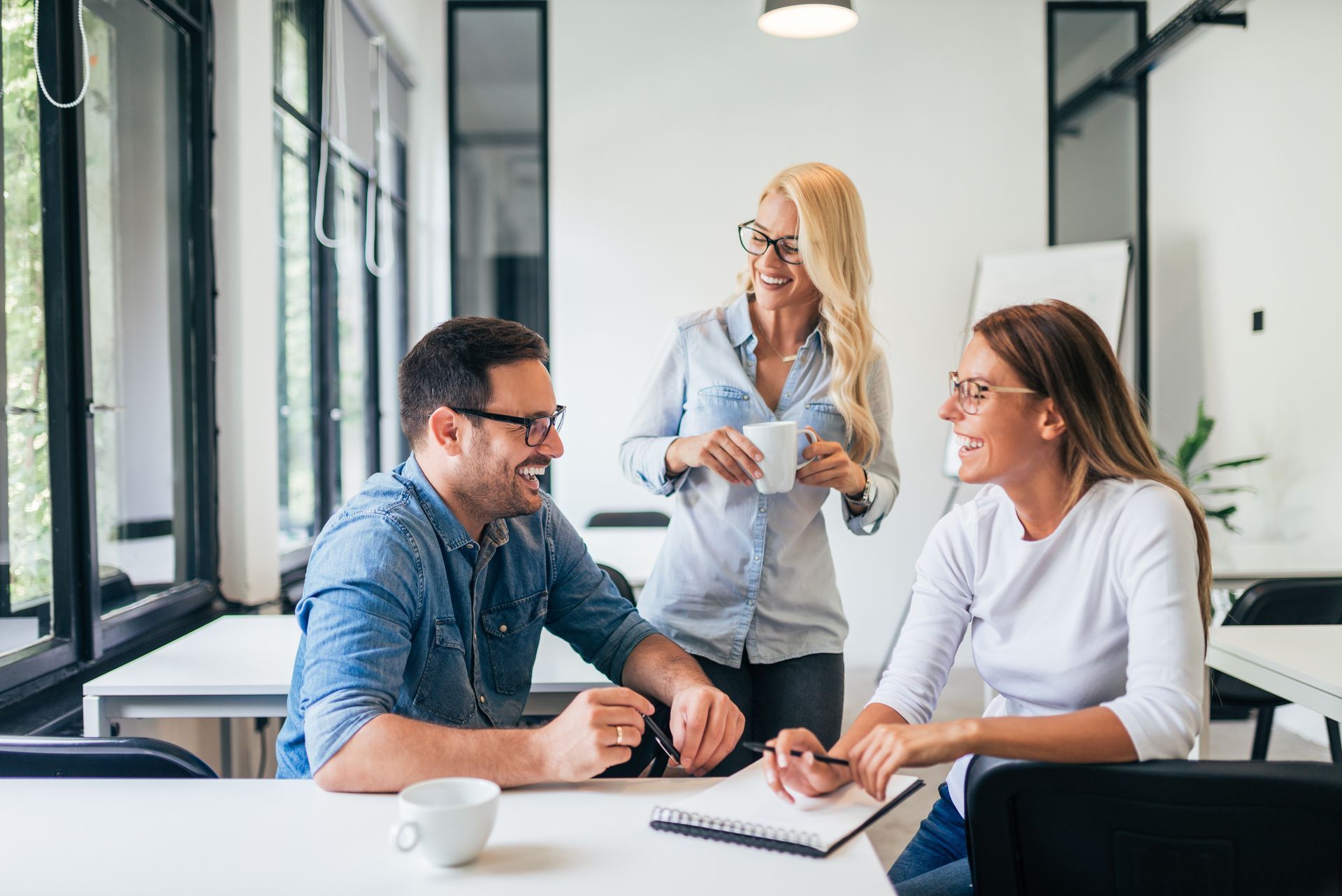 Three colleagues in a bright office laughing and collaborating over a notebook on a desk.