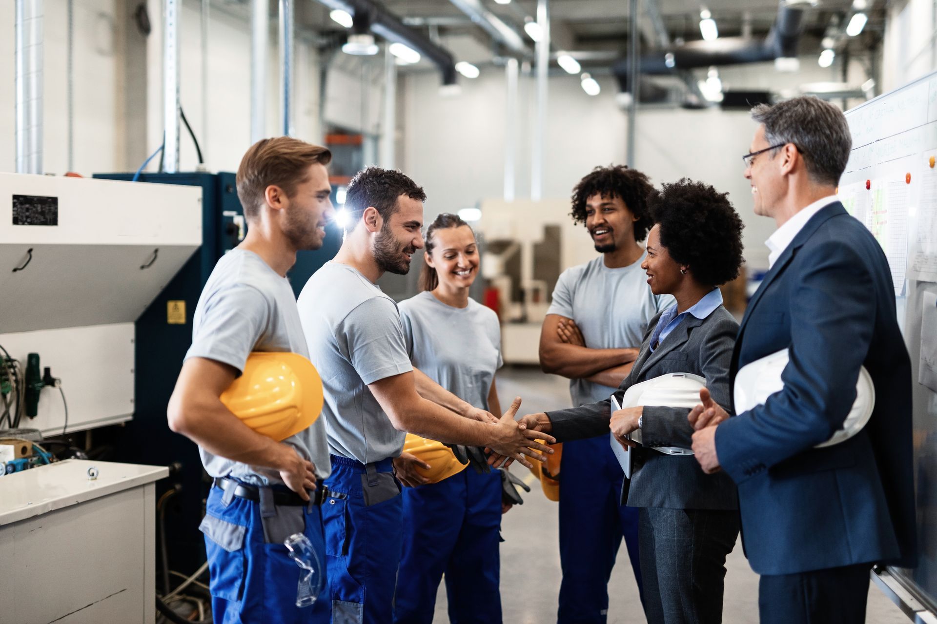 A professional team shakes hands in a factory setting while colleagues look on and smile.