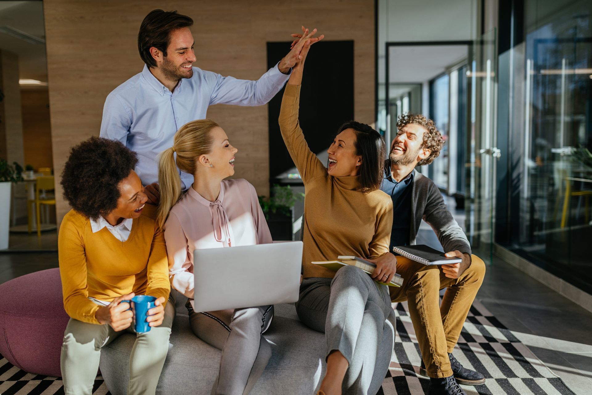 A diverse group of colleagues in an office workspace are laughing and giving a high-five while using a laptop.