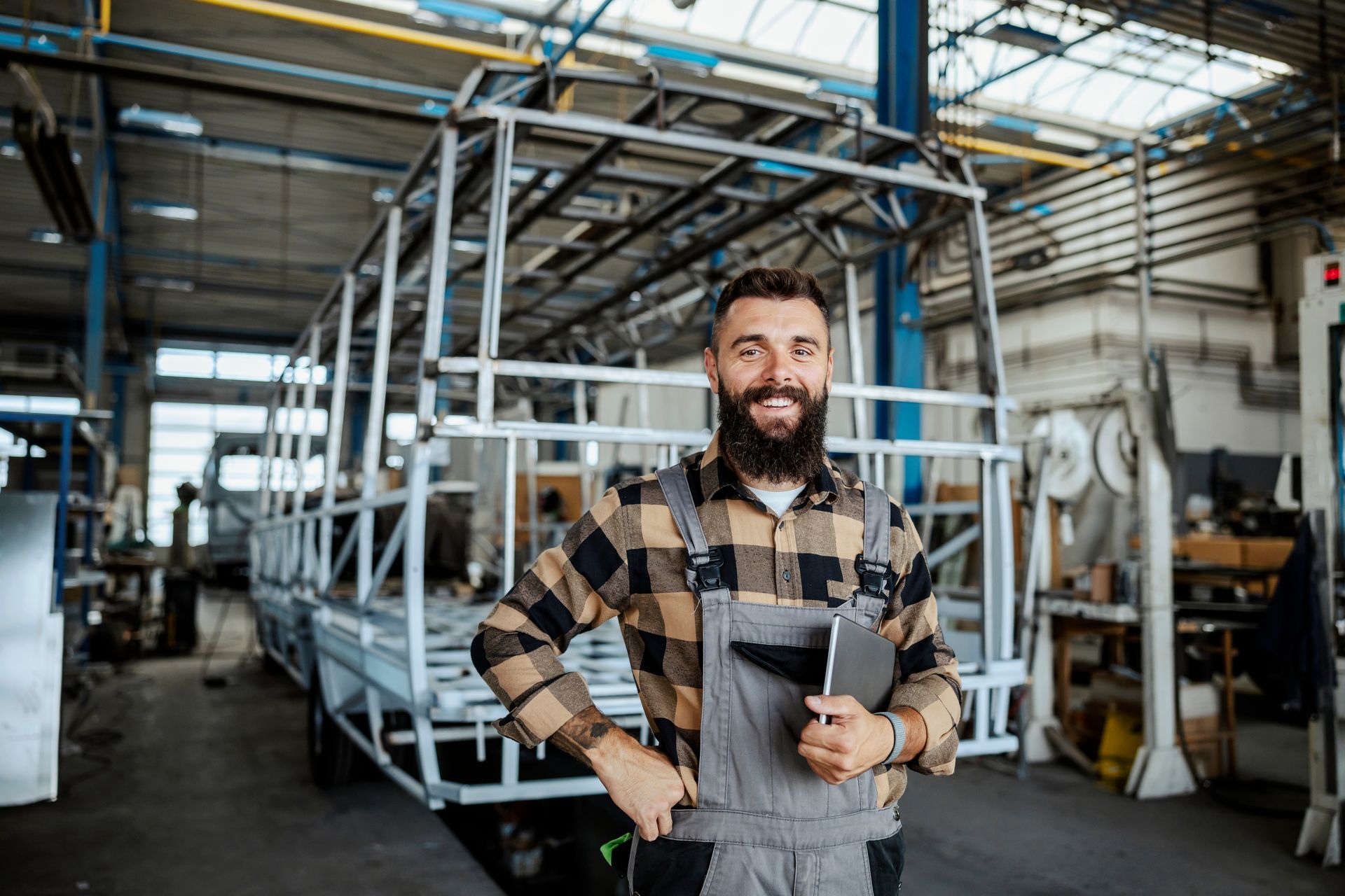 A smiling worker in a flannel shirt and overalls holds a tablet in front of a metal vehicle frame in a factory.