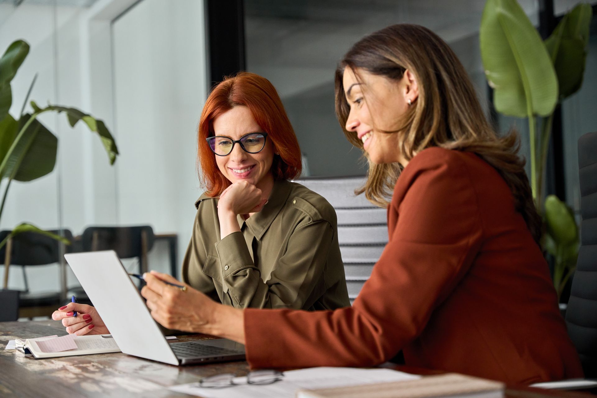 Two colleagues at a wooden desk look at a laptop screen, smiling as they discuss their work in a bright, modern office.