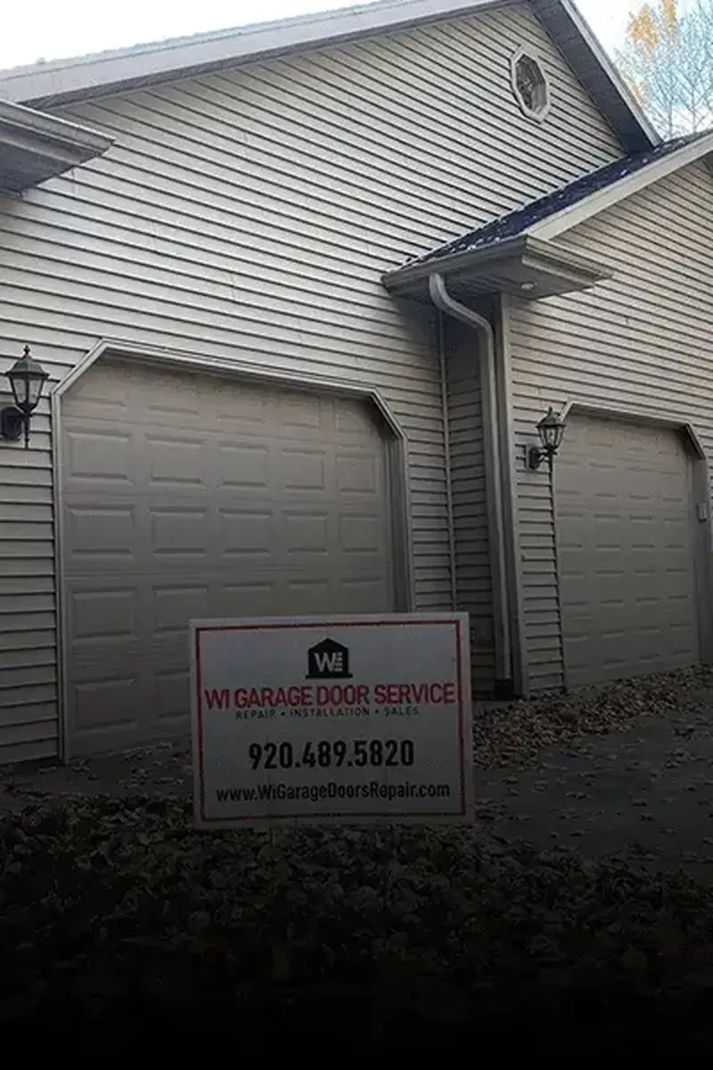 A business sign for WI Garage Door Service stands in front of a house with two beige garage doors and exterior lights.