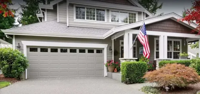 A gray two-story house with a two-car garage, covered porch, and an American flag near the front entrance.