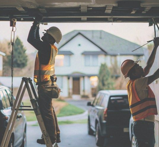 Two workers in safety vests and hard hats install equipment in a garage with a house and truck outside