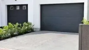 A modern residential driveway with two dark grey garage doors and a row of green bushes in front.