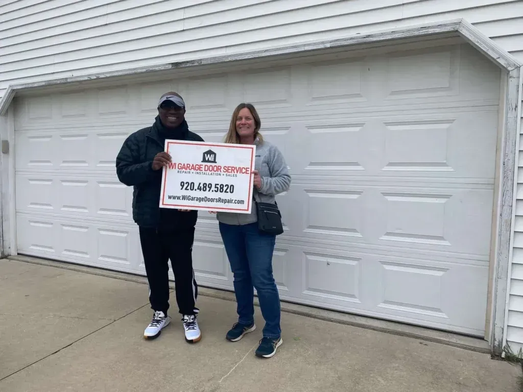 Two people stand before a white residential garage door holding a sign for 