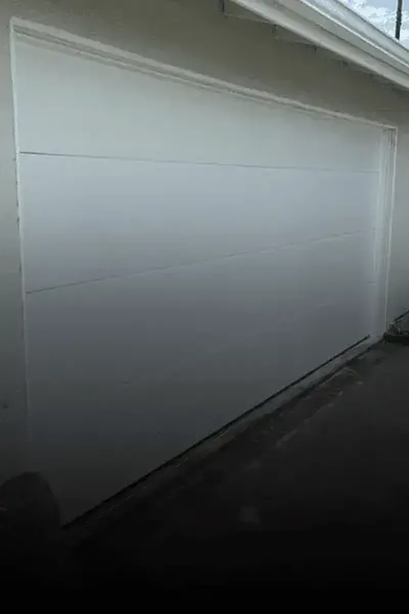A plain, white, multi-panel garage door viewed from an angle against a concrete exterior wall.