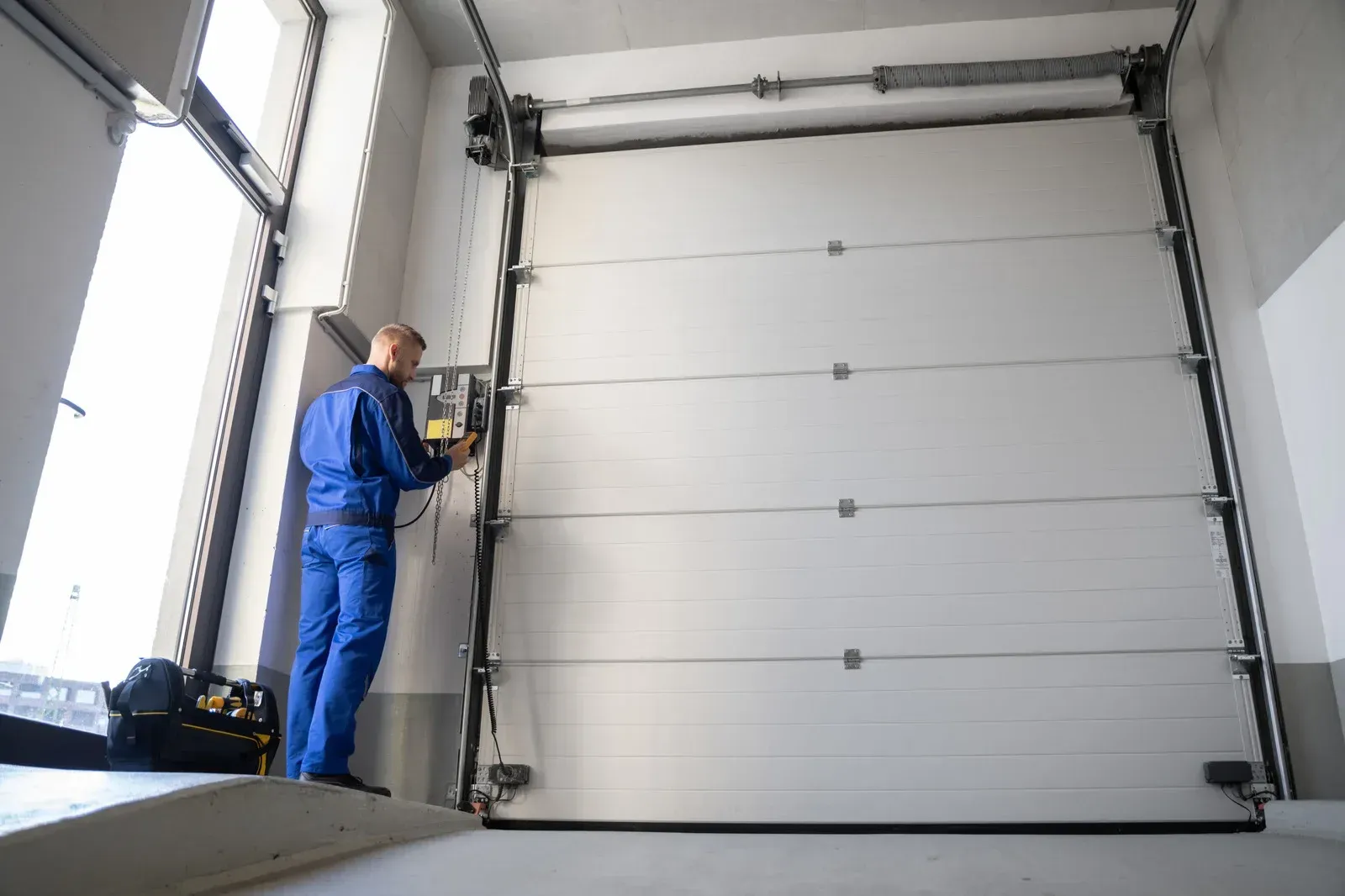 Worker in blue coveralls inspecting a large white industrial garage door inside a warehouse