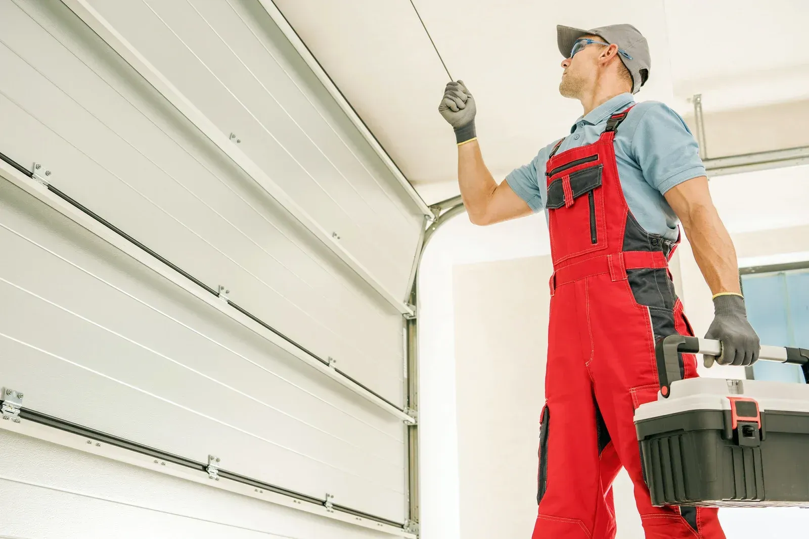 Technician in red overalls inspects a garage door track while holding a toolbox.