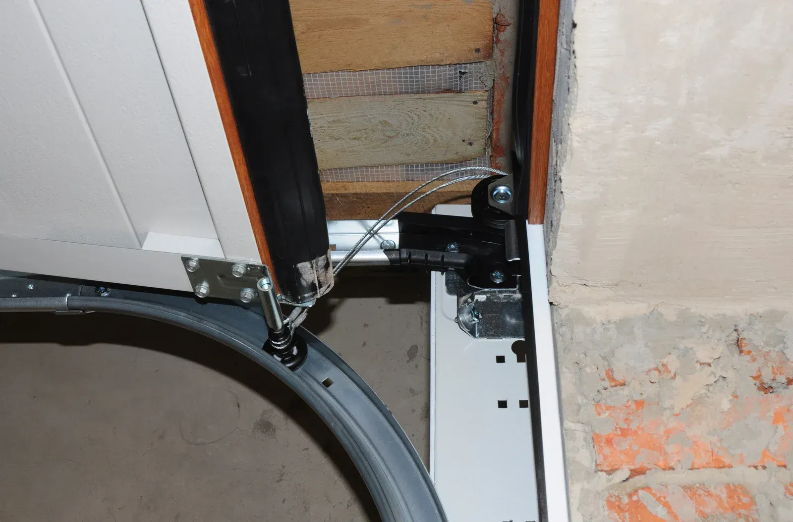 Close-up of a garage door track and hinge beside a wall, with a curved rubber seal on the floor.