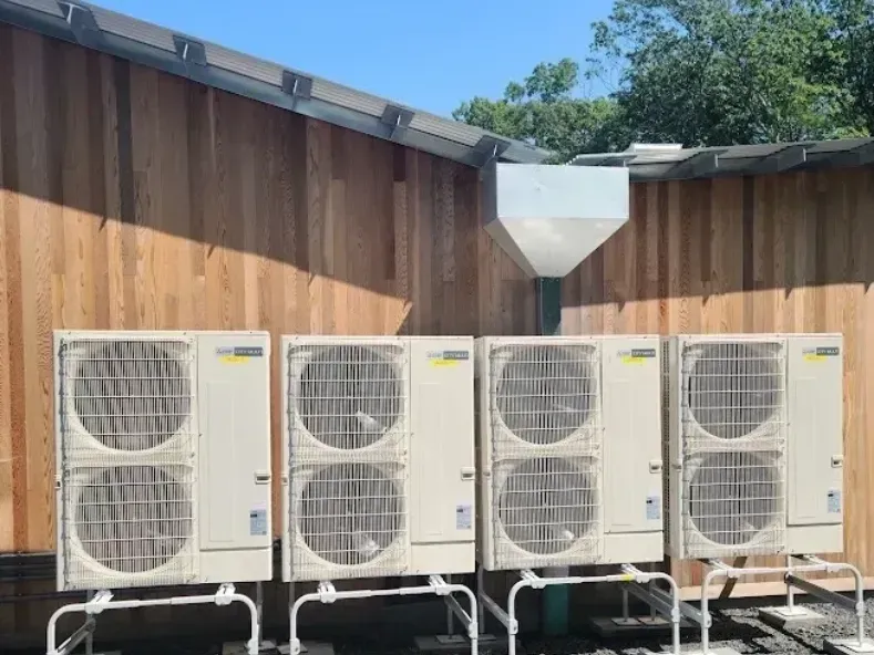 Four cream-colored outdoor air conditioning units stand in a row against a light wood-paneled wall.