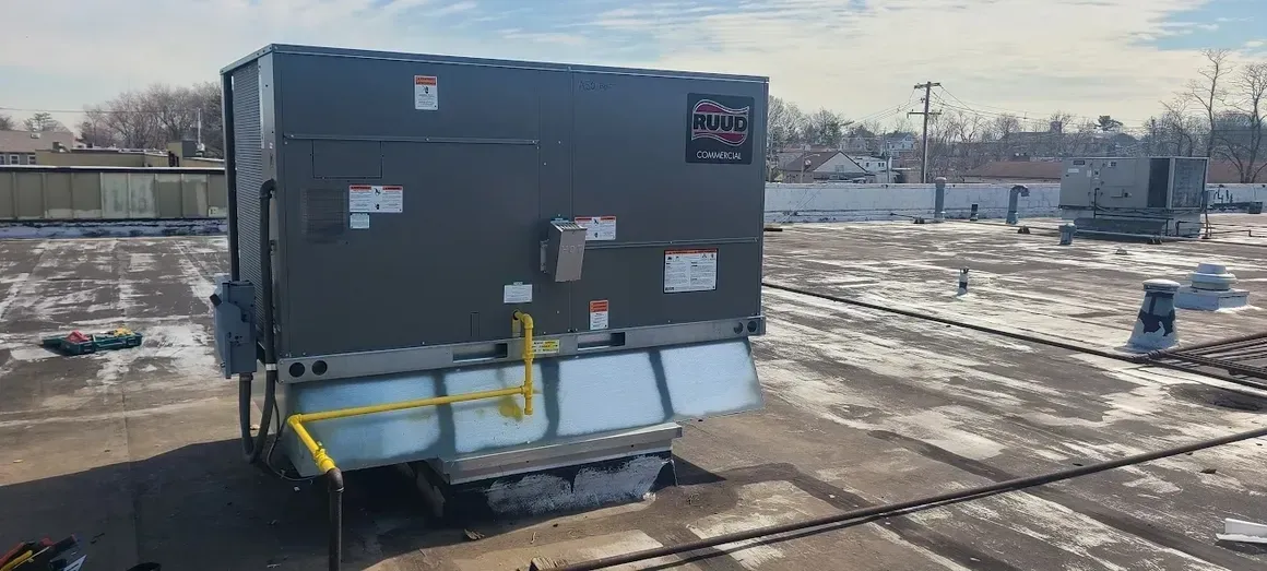 A gray commercial HVAC rooftop unit with yellow gas piping, situated on a snowy flat roof under a bright sky.