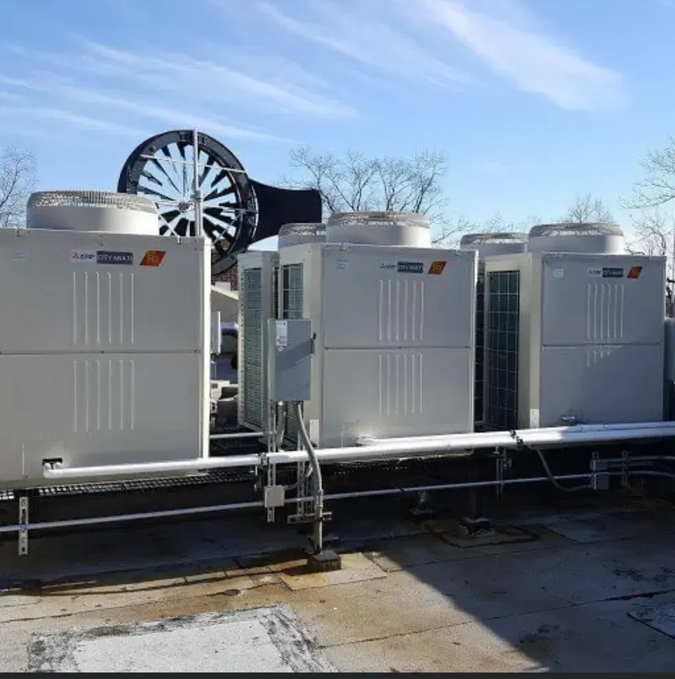 Three industrial HVAC units on a flat roof under a blue sky, with a large, dark circular ventilation fan in the background.