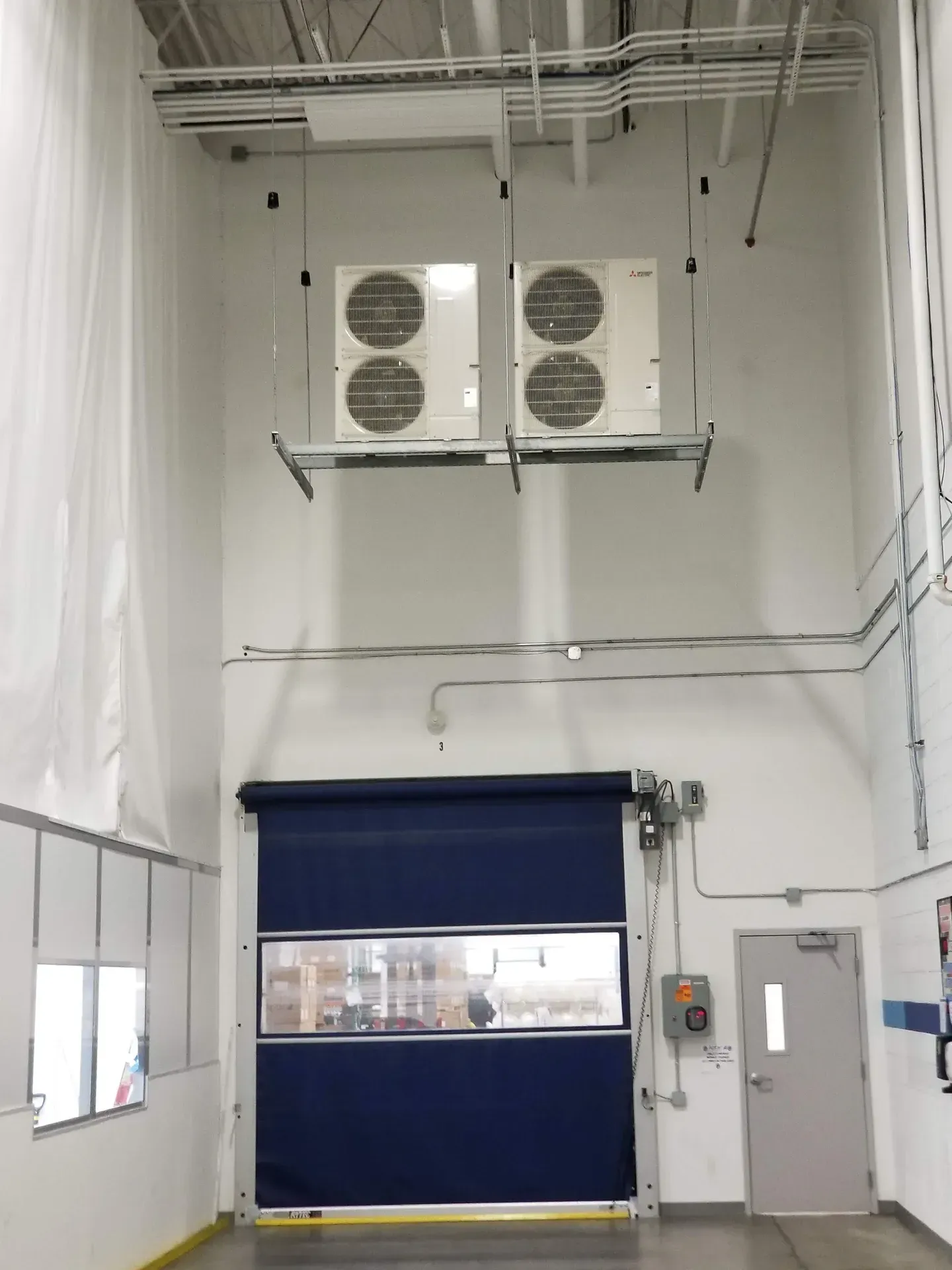 A high-ceiling industrial room with two suspended HVAC units, a large blue roll-up door, and a gray side door.
