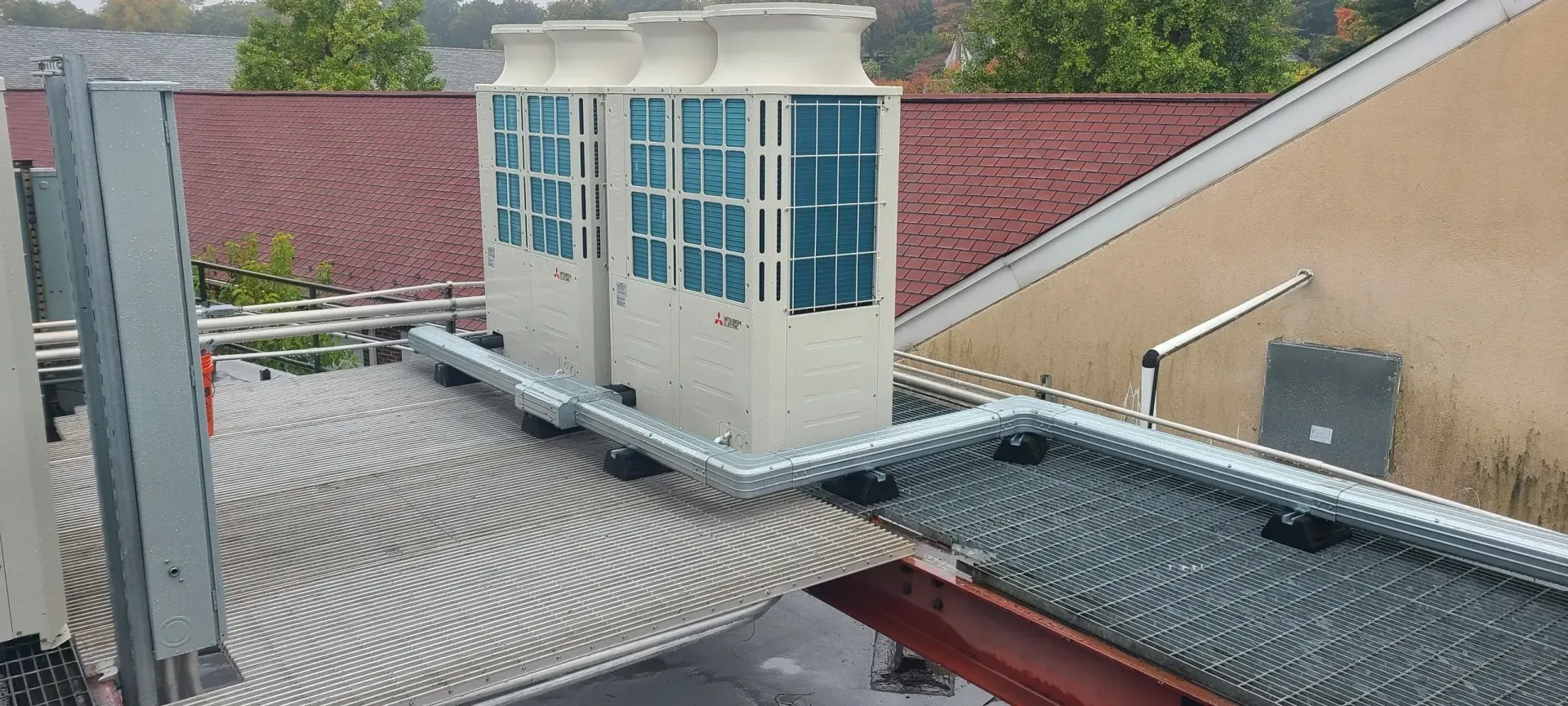 Two HVAC units sit on a roof with a patterned surface, with metal bird spikes installed along the pipes and nearby edges.