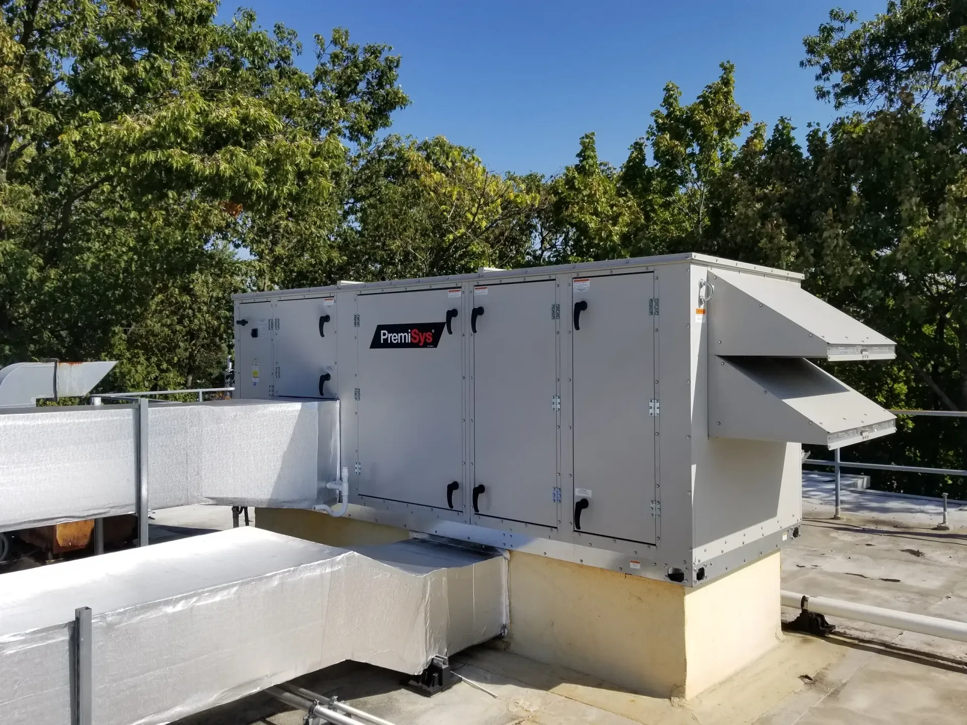 A light gray commercial HVAC rooftop unit with metal ductwork stands on a flat roof against a background of green trees.