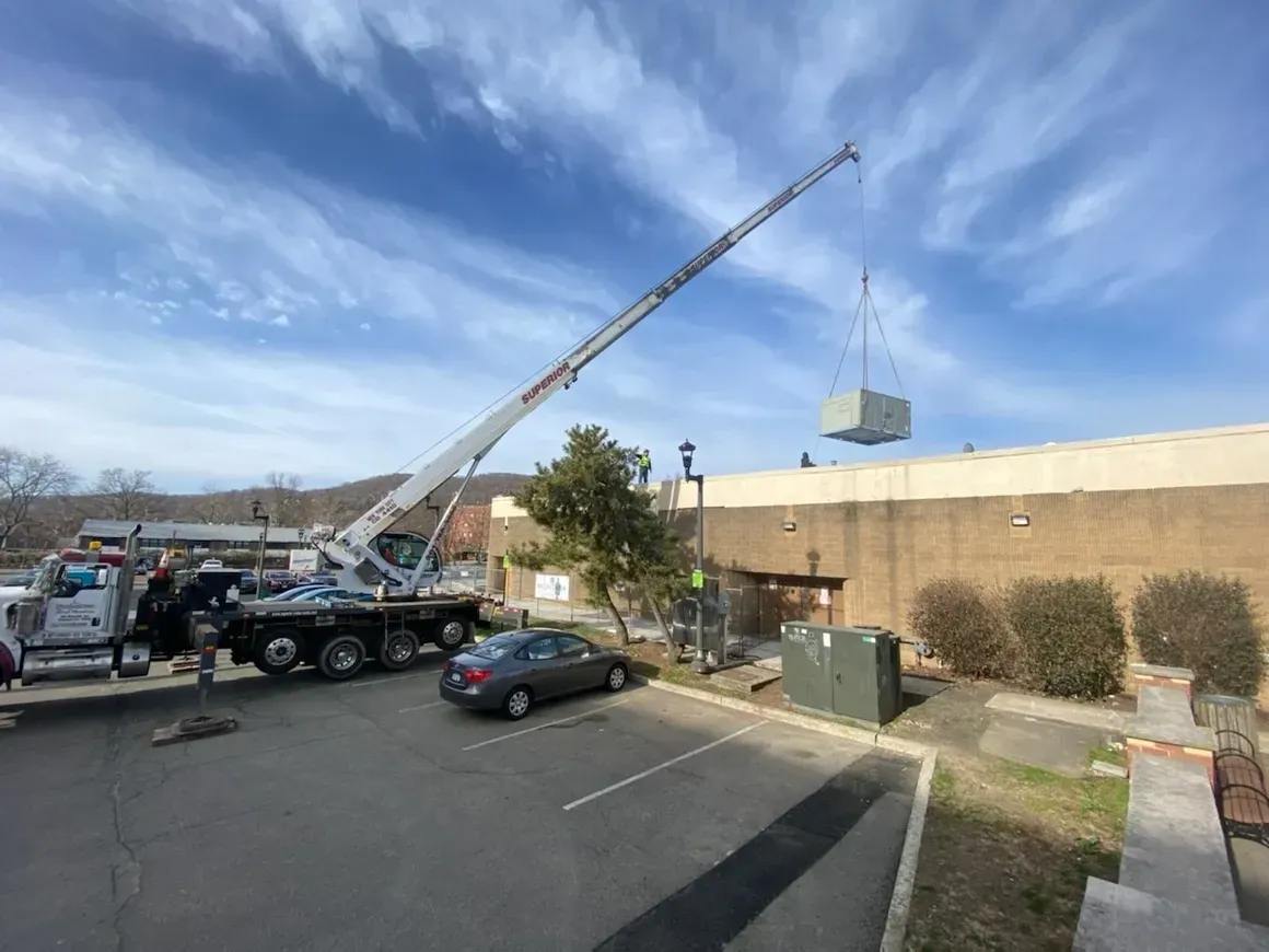 A mobile crane hoists a large HVAC unit above a commercial building rooftop in a sunny parking lot.