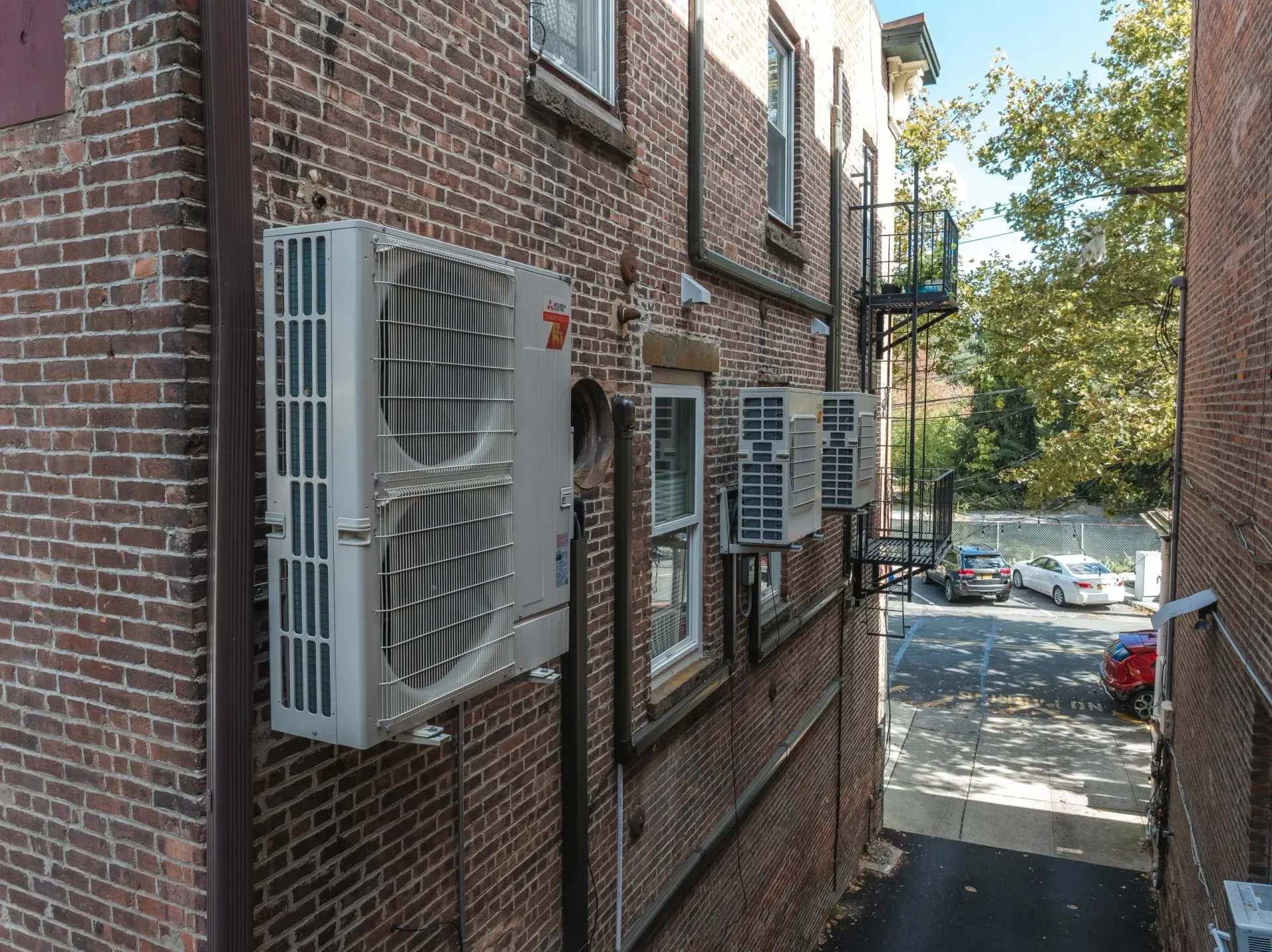 Three air conditioning units are mounted on a brick building wall, overlooking a parking lot with several cars.
