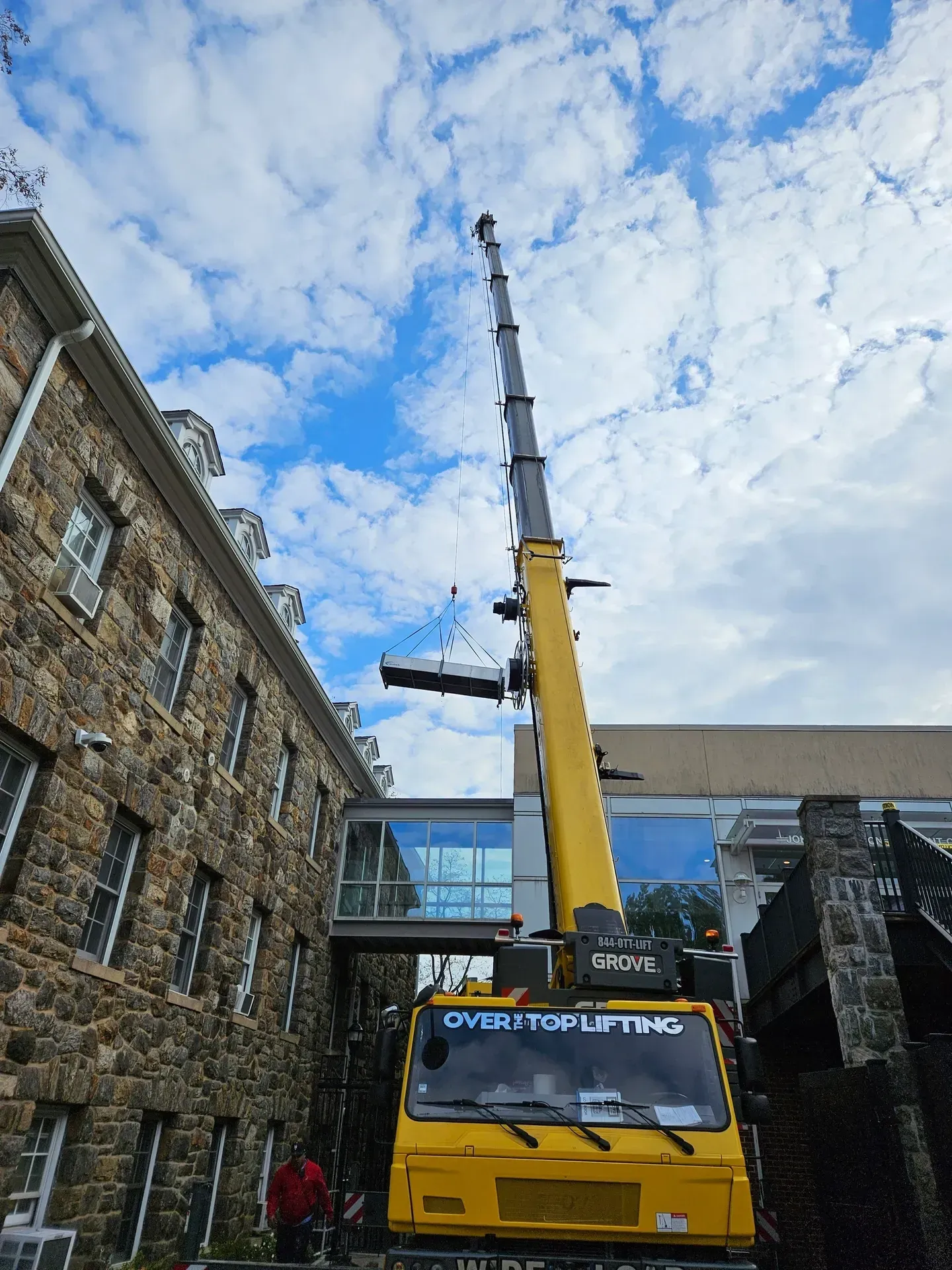 A yellow crane lifting a large rectangular object toward a glass skybridge between a stone building and a modern structure.