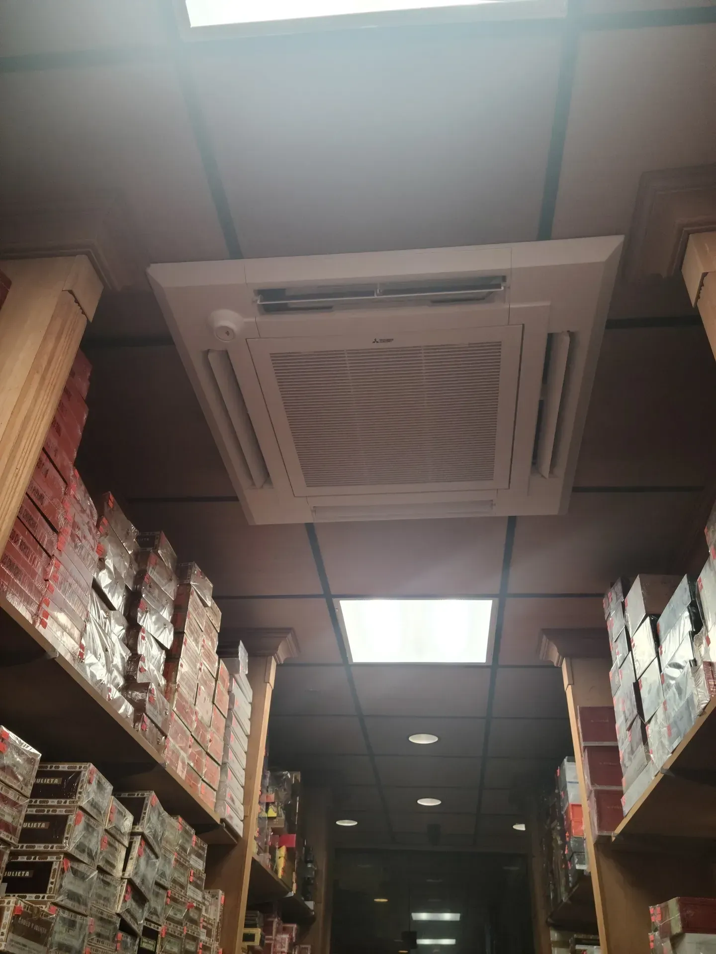 A ceiling-mounted air conditioning unit in a narrow aisle between tall shelves stocked with boxes in a retail store.