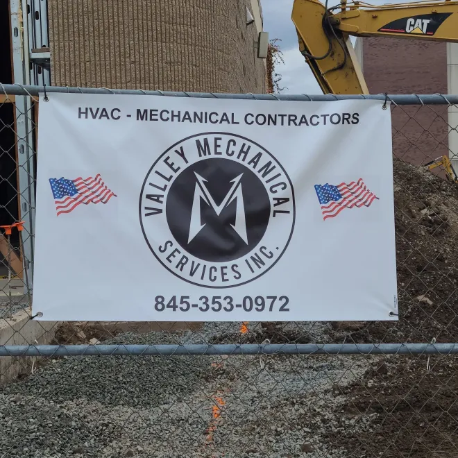 A banner for Valley Mechanical Services, Inc. featuring their logo, contact number, and American flags on a chain-link fence.