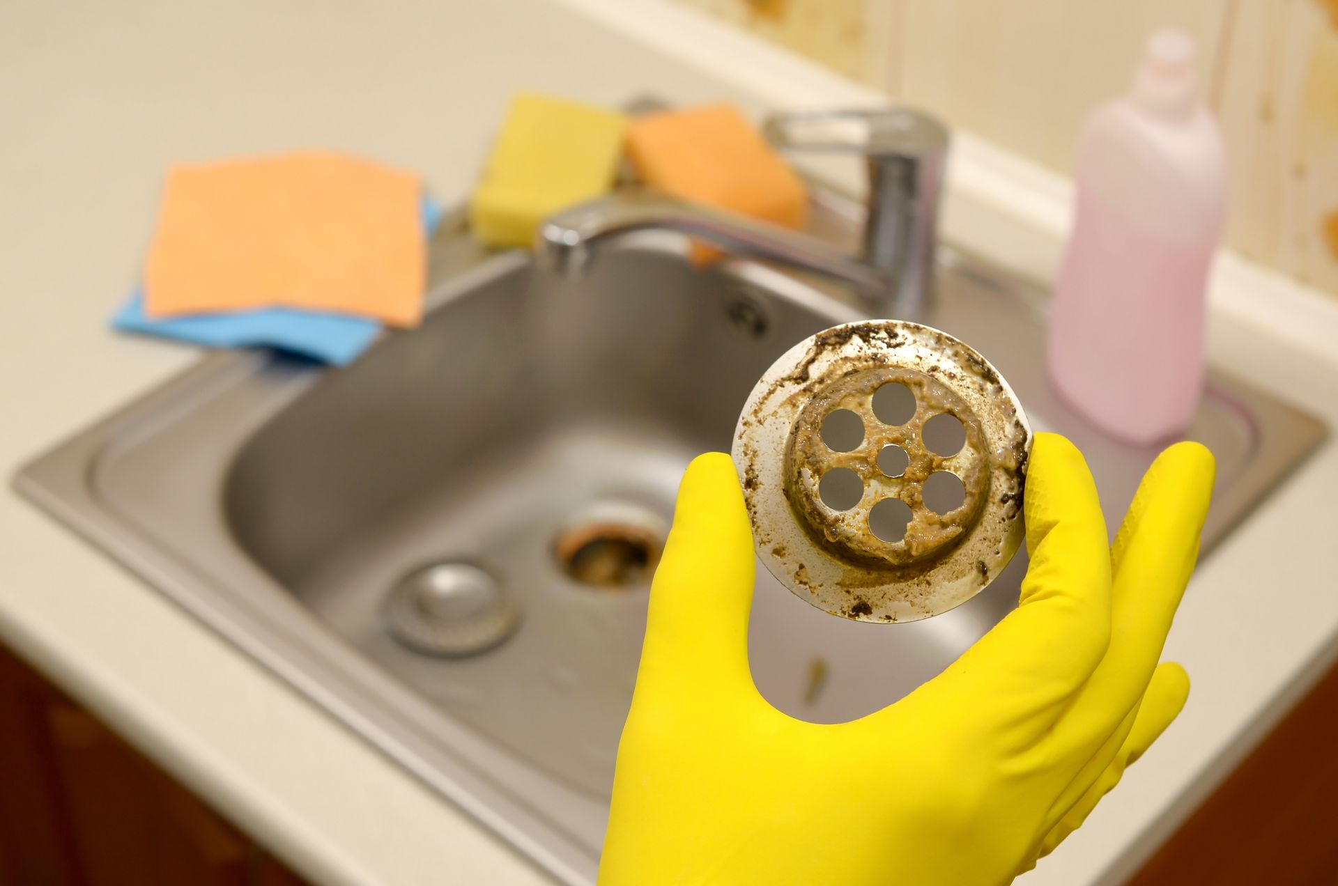 Yellow-gloved hand holding a dirty sink drain strainer over a kitchen sink