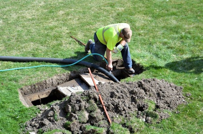 Worker in a neon vest digging near an open ground hole beside hoses on a grassy lawn