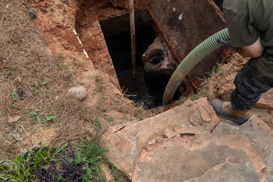 Person lowering a hose into a narrow dirt pit beside a rocky edge