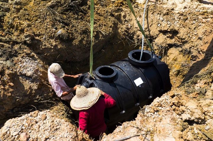 Workers install a large black underground tank in a dirt excavation pit.