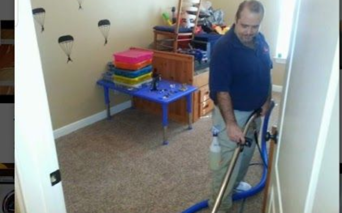 A person in rain gear uses a yellow industrial floor scrubber to clean a soapy, wet rug.