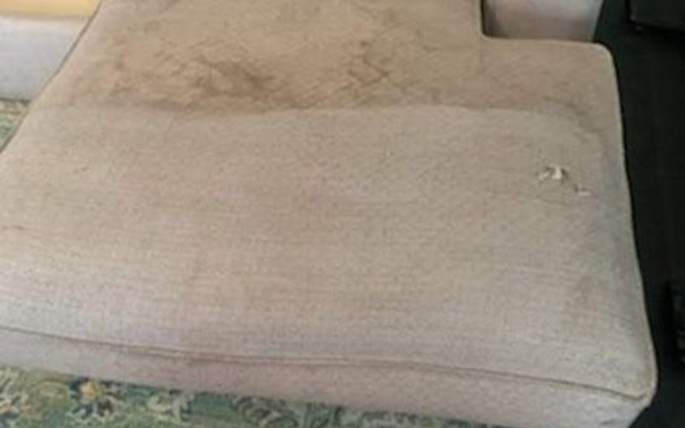 A person wearing a blue apron and yellow gloves vacuums a light-colored rug in a living room with a sofa and ottoman.
