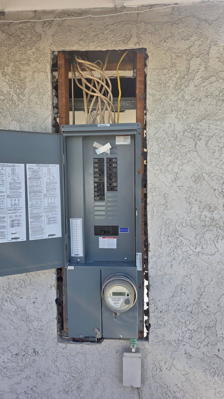 Open electrical panel in stucco wall with exposed wiring above and utility meter below.