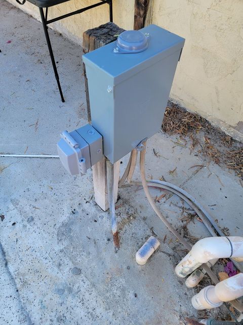 Gray electrical box on a post beside concrete walkway with pipes and cords nearby