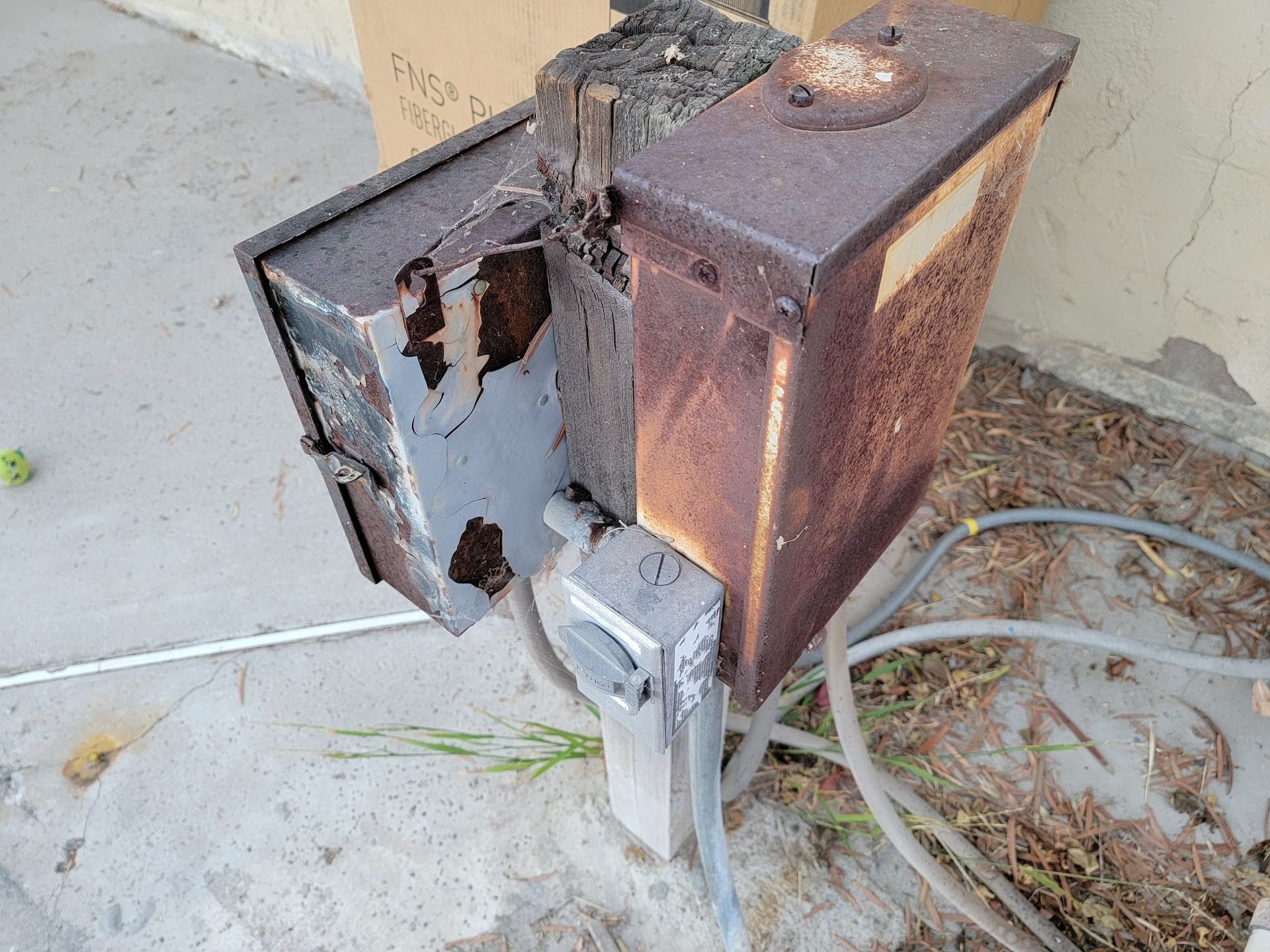 Rusty outdoor electrical box with an open door and cables on the ground against a wall