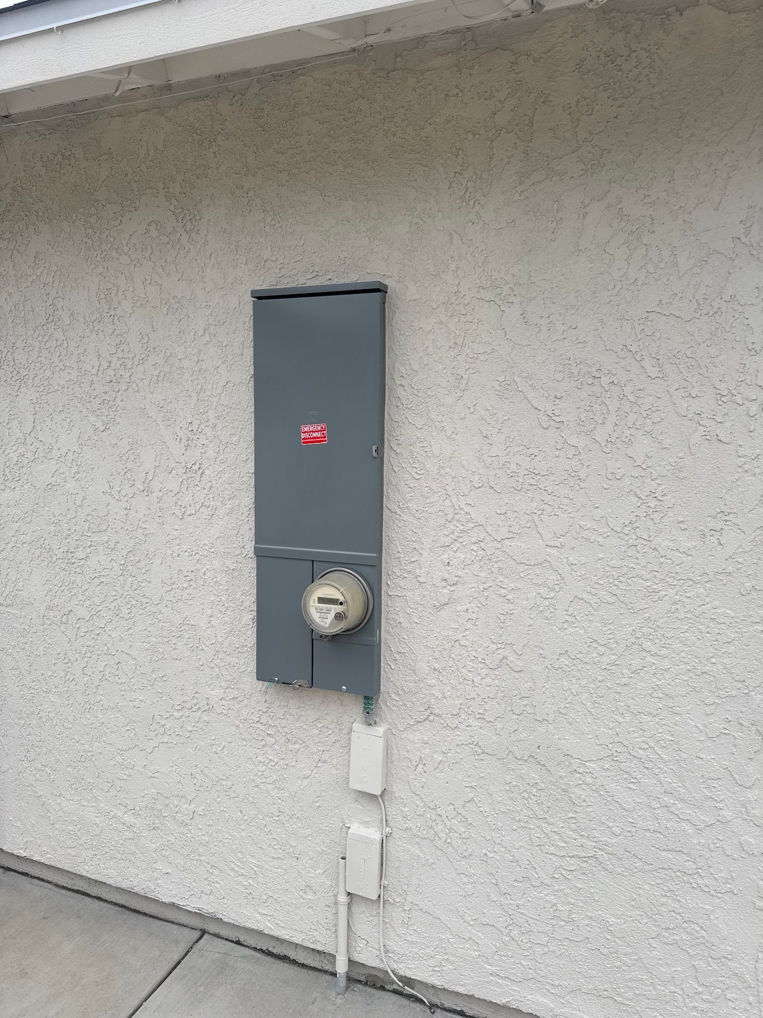 Gray electrical meter box mounted on a stucco exterior wall with conduit below.