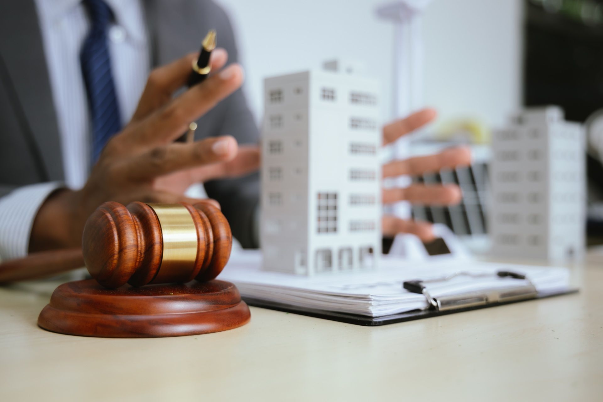 A person in a suit gestures towards model buildings, with a gavel on a desk.