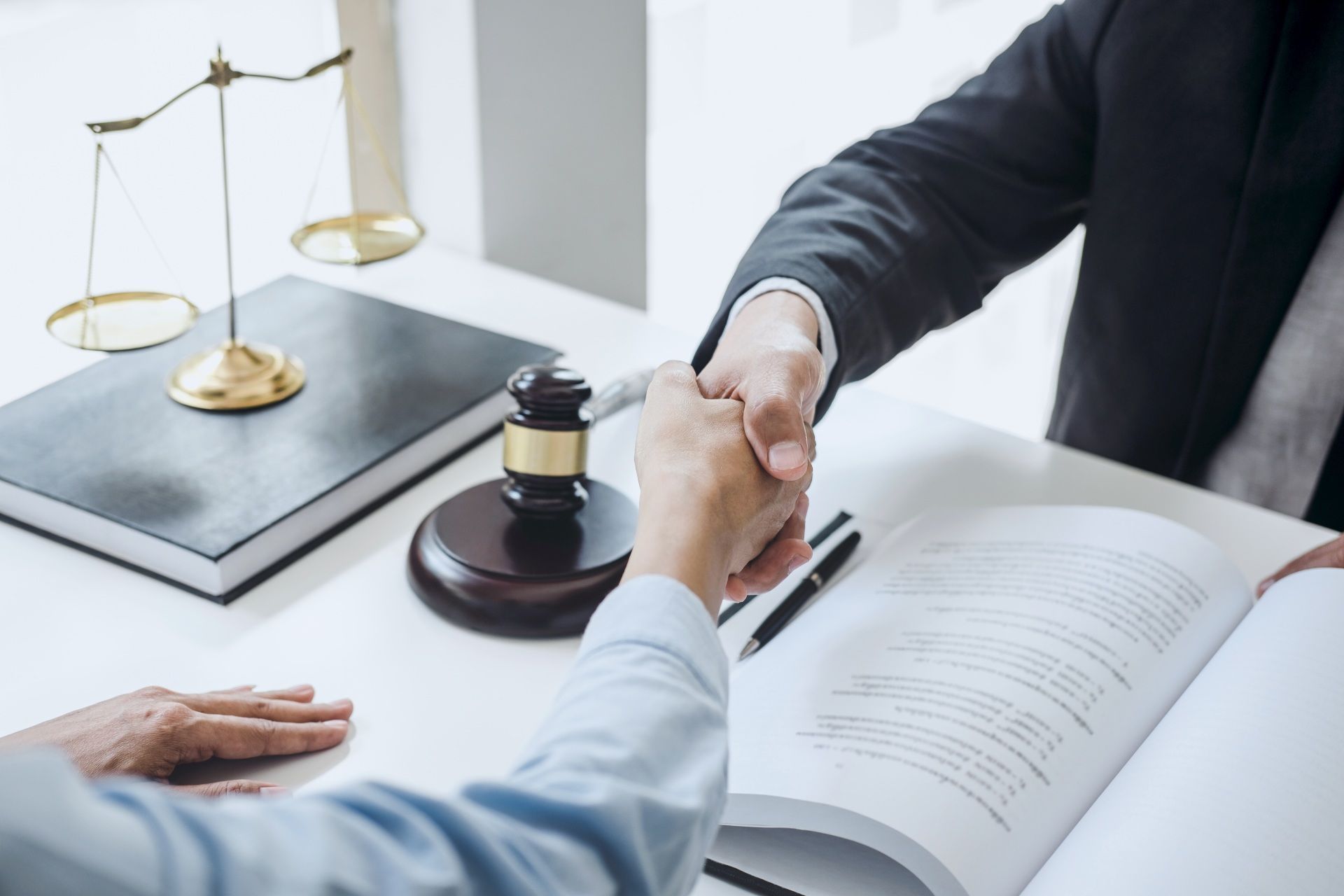 Two people shaking hands at a desk with a gavel, scales of justice, and documents.